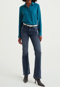 Turquoise langarmige shirt gecombineerd met donkerblauwe jeans en een witte riem. De jeans hebben een rechte pijp en de outfit wordt aangevuld met een paar grijze hoge hakken.
