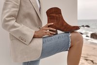 Personne portant un blazer beige et un short en jean déchiré tient une botte cheville en daim marron sur un genou plié, au bord de la plage avec des vagues et des rochers en arrière-plan.