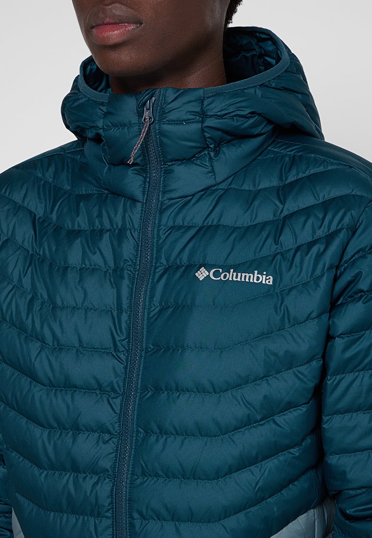 Chaqueta acolchada color verde azulado con capucha, con diseño acolchado, cierre de cremallera y logo de Columbia en el lado izquierdo del pecho. Textura lisa y ligera.