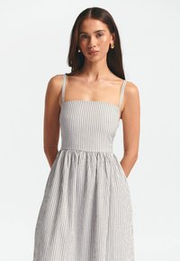 Bezlankstė suknelė su vertikaliomis tamsiai mėlynomis ir baltomis juostomis, turinti prigludusią liemenę ir laisvai krentantį, susiraukšlėjusį sijoną, pagaminta iš lengvos medžiagos.