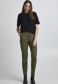 Zwart gestructureerd T-shirt met korte mouwen en verhoogde ribbels, gecombineerd met olijfgroene slim-fit broek en zwarte enkelboots. Eenvoudige, moderne outfit.