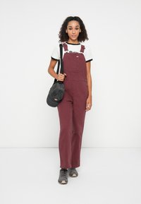 Une salopette bourgogne en coton avec une poche avant, portée sur un T-shirt rayé noir et blanc ; associée à des chaussures grises et un sac noir.