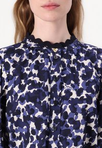 Bluse mit floralem Muster in Blau und Creme, mit einem gewellten Kragen, goldenen Herzknopf-Akzenten und schwarzen Knöpfen an der Vorderseite.