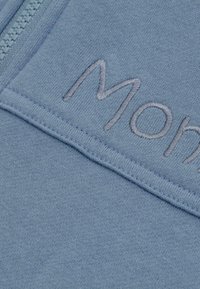 Blauer Sweatshirt mit weicher Textur, ausgestattet mit einem gestickten "Mon"-Design in Hellgrau in der Nähe des Reißverschlusses. Sichtbare Nähte.