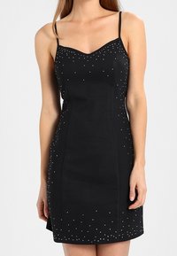 Svart miniklänning med tunna band, med en figurnära design och dekorerad med spridda metallknappar längs bysten och fållen.