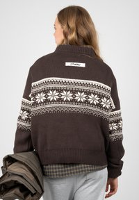 Femme portant un pull marron foncé avec un motif flocon de neige blanc, tenant une veste marron pliée, de dos sur un fond uni.