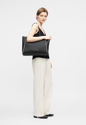 Sieviete stāv sānis, valkājot melnu piedurkņu topu, baltas platām kājām bikses, melnas kurpes, plecā turot lielu melnu Love Moschino somu.