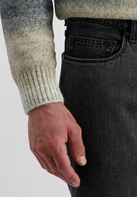 Brede trui met een verloop van groen naar beige, met geribbelde manchetten. Gecombineerd met donkere spijkerbroek. Focus op hand die naast de zak rust.