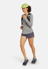 Graues Langarmshirt, dunkle Shorts, schwarze Laufschuhe mit lila Akzenten und eine leuchtend grüne Kappe; trägt einen Trinkrucksack.