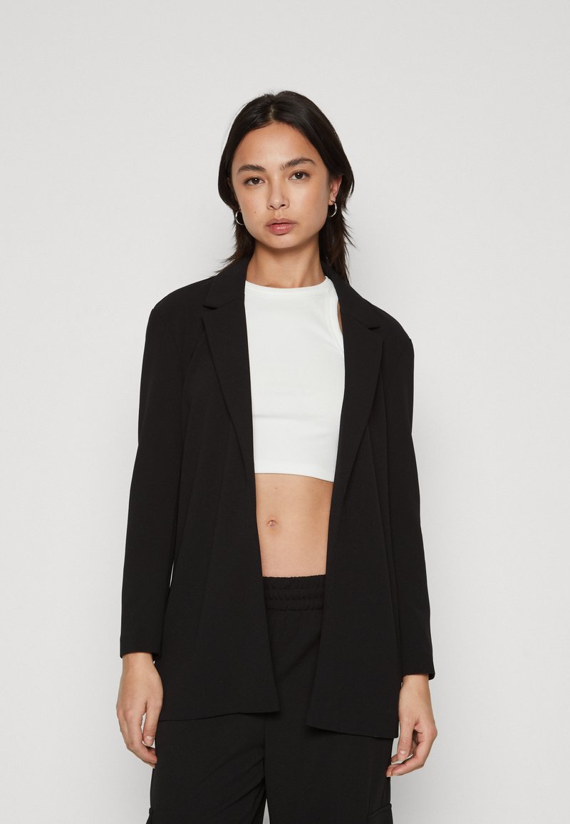 Fekete blézer testreszabott kialakítással, kivágott gallérral és hosszú ujjal, fehér crop top fölött viselve, sima textúrával.