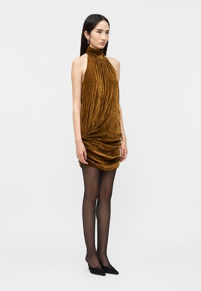 Ženska, oblečena v brezrokavno mini obleko iz rjavega žameta z visokim ovratnikom, črnimi prosojnimi nogavicami in črnimi čevlji z ostrimi konicami, stoji ob preprostem ozadju.