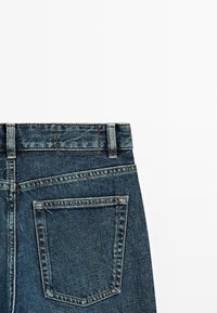 Granatowe dżinsy o średnim stanie, z szlufkami na pasek i jedną tylną kieszenią z kontrastowym przeszyciem. Gładka tekstura.