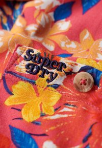 Un tissu à motif floral dans des tons vifs d'orange, de jaune et de bleu présente une étiquette brodée "Superdry" et un bouton crème avec un détail floral.