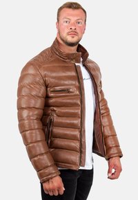 Giubbotto imbottito in pelle marrone con collo alto, zip frontale e due tasche con zip. Presenta una texture a righe verticali e polsini a coste.