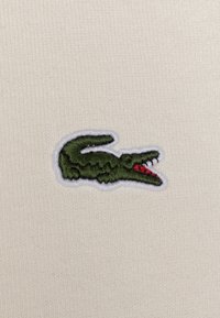 Logotipo de cocodrilo bordado en verde con acentos rojos sobre un fondo de tela ribeteada crema. Texturizado, con un borde blanco contrastante.