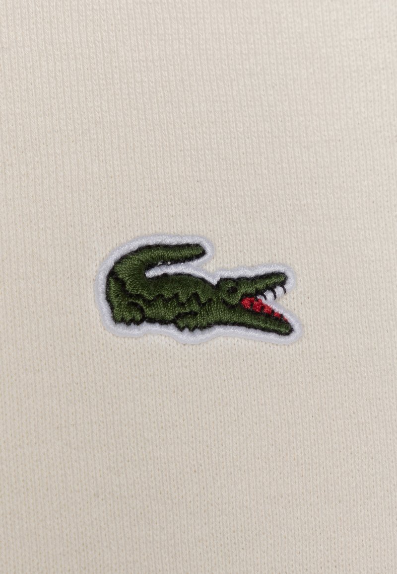 Logo di coccodrillo verde ricamato con accenti rossi su uno sfondo di tessuto a coste color crema. Testurizzato, con un bordo bianco a contrasto.