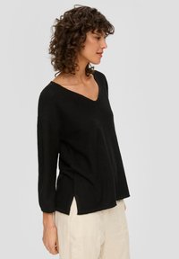 Schwarzer Strickpullover mit V-Neck, herunterfallenden Schultern und seitlichen Schlitzen. Weiche Textur, lockere Passform. Kombiniert mit hellen Hosen.