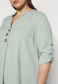 Blouse vert clair avec un décolleté en V et trois boutons décoratifs. Texture douce, manches retroussées maintenues par des accents de bouton, coupe ample.