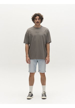 Kohalike krussis juuste ja vuntsidega mees seisab otsevaates, seljas üle suurusega hall T-särk, heledad teksashortsid, valged sokid ja mustad tossud.