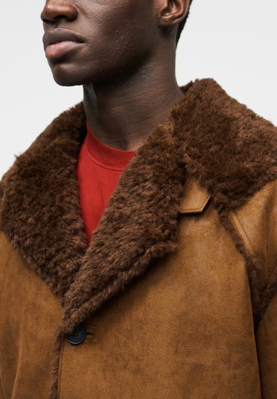 Καφέ παλτό από ψεύτικο σουέτ με πλούσιο καφέ γούνινο γιακά, με κούμπωμα κουμπιών και λεία υφή, φορεμένο πάνω από κόκκινο πουκάμισο.