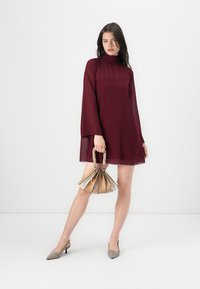 Dieprode lange jurk met een hoge hals en vloeiende stof. Model houdt een geplooide, beige handtas vast en draagt taupe hakken.