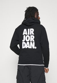 Felpa nera con cappuccio e grande logo bianco "AIR JORDAN" sul retro. Presenta un cappuccio regolabile e polsini a costine, abbinata a pantaloni cargo scuri.