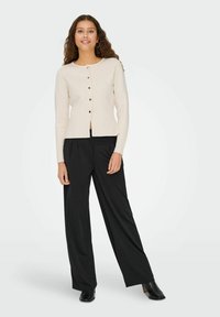 Cardigan créme à col rond, avec des boutons noirs et une coupe droite, associé à un pantalon large noir et des bottines à talons noires.