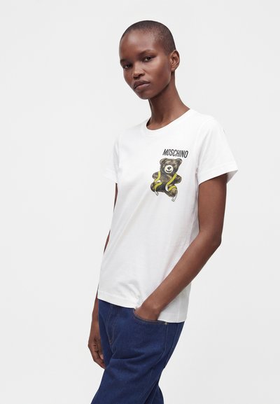 Άτομο που φοράει ένα λευκό t-shirt Moschino με γραφικό teddy bear και μπλε τζιν, στέκεται με το ένα χέρι στην τσέπη μπροστά σε απλό φόντο.