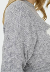 Pull en tricot gris avec un motif texturé, présentant un col montant et des coutures visibles. Une partie du design comprend un accent de couleur claire.