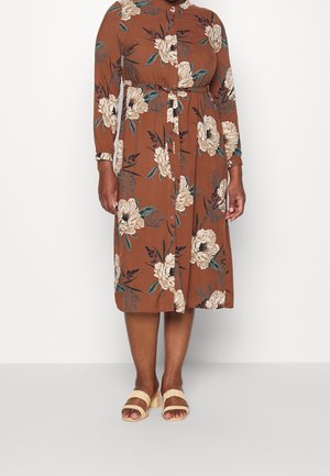 Woman wearing a brown, long-sleeve, buttoned dress with large white floral prints and beige heeled sandals standing on a plain background.