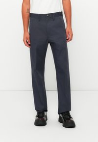 Uomo che indossa pantaloni dritti di un blu scuro, stivali neri robusti e una maglietta bianca su uno sfondo bianco.