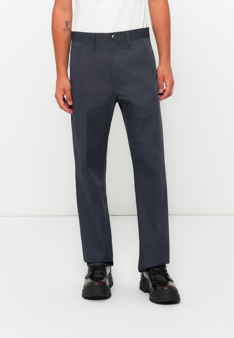 Uomo che indossa pantaloni dritti di un blu scuro, stivali neri robusti e una maglietta bianca su uno sfondo bianco.