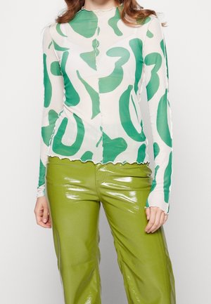 Vrouw die een wit shirt met lange mouwen draagt met groene abstracte vormen en glanzende groene hoge taille broek.