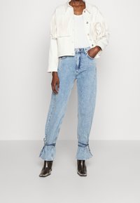 Chaqueta blanca con flecos sobre una blusa blanca, combinada con unos vaqueros de cintura alta en azul claro con puños atados y botas negras de tacón.