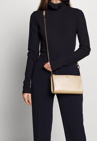 Złota skórzana torebka na łańcuszku. Posiada gładką fakturę i prostokątny kształt. Czarna, prążkowana bluzka z długim rękawem oraz taliowane spodnie.