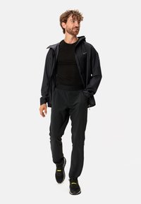 Giacca nera impermeabile con cappuccio, abbinata a una maglietta nera a maniche lunghe aderente e pantaloni neri leggeri. Presenta dettagli gialli nelle scarpe.