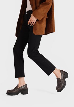 Mocassini in pelle marrone con tacco, caratterizzati da una finitura testurizzata e una sottile decorazione orizzontale; indossati con jeans neri a vita alta e un blazer in suede marrone.