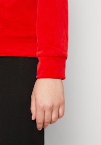 Hände an der Seite, trägt ein rotes Langarmoberteil und schwarze Hosen, vor einem schlichten hellgrauen Hintergrund.