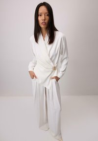 Bel top z belo prevezo in široke hlače iz gladkega blaga. Top ima dekorativen zlat poudarek; oba kosa imata čist, minimalističen dizajn.