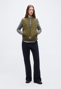 Γυναίκα που στέκεται με τα χέρια στις τσέπες φορώντας ένα πράσινο ελιάς γιλέκο με καπιτονέ ραφές, γκρι πουλόβερ, ριγέ πουκάμισο, σκοτεινό τζιν και καφέ παπούτσια.