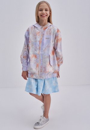 Kurtka tie-dye w pastelowych odcieniach fioletu i pomarańczu, z zamkiem z przodu, dwoma kieszeniami i elastycznymi mankietami. Nosi się ją na jasnoniebieskiej spódnicy.