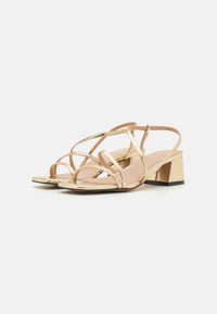 Sandales à talons métalliques dorées avec plusieurs sangles croisées, un talon carré et une texture accentuée au niveau du talon.