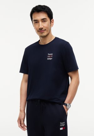 Homme portant un t-shirt Tommy Hilfiger bleu marine et un pantalon assorti, avec des logos sur la poitrine et la cuisse, debout avec une main dans la poche.