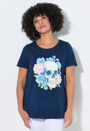Donna con capelli ricci che indossa orecchini a cerchio e una maglietta blu navy con un teschio bianco circondato da fiori blu e rosa.