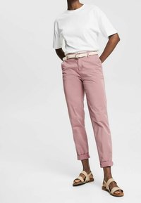 Persoon in een wit T-shirt met korte mouwen, roze opgerolde broek met een gevlochten riem en beige sandalen met open tenen, staande met de handen in de zakken.