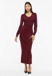 Vestido de punto burdeos de manga larga con escote redondo, silueta ajustada y detalles de hombros ligeramente abullonados, combinado con tacones altos negros.