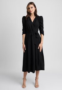 Kobieta stoi w czarnej midi sukience typu wrap z bufiastymi ramionami i beżowych sandałach na kostkach na jednolitym szarym tle.