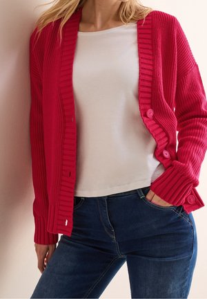 Magenta ribbed cardigan with buttons, worn over a white shirt. Features a relaxed fit and textured sleeves, paired with blue jeans.