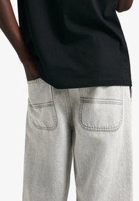 Fekete pamut póló világosszürke farmernadrággal párosítva. A nadrág két hátsó zsebbel rendelkezik, kontrasztos varrással és strukturált felülettel.