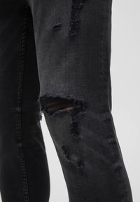 Svarta denimjeans med flera slitningar och revor, inklusive ett stort hål som blottar huden på låret, visat mot en enfärgad bakgrund.